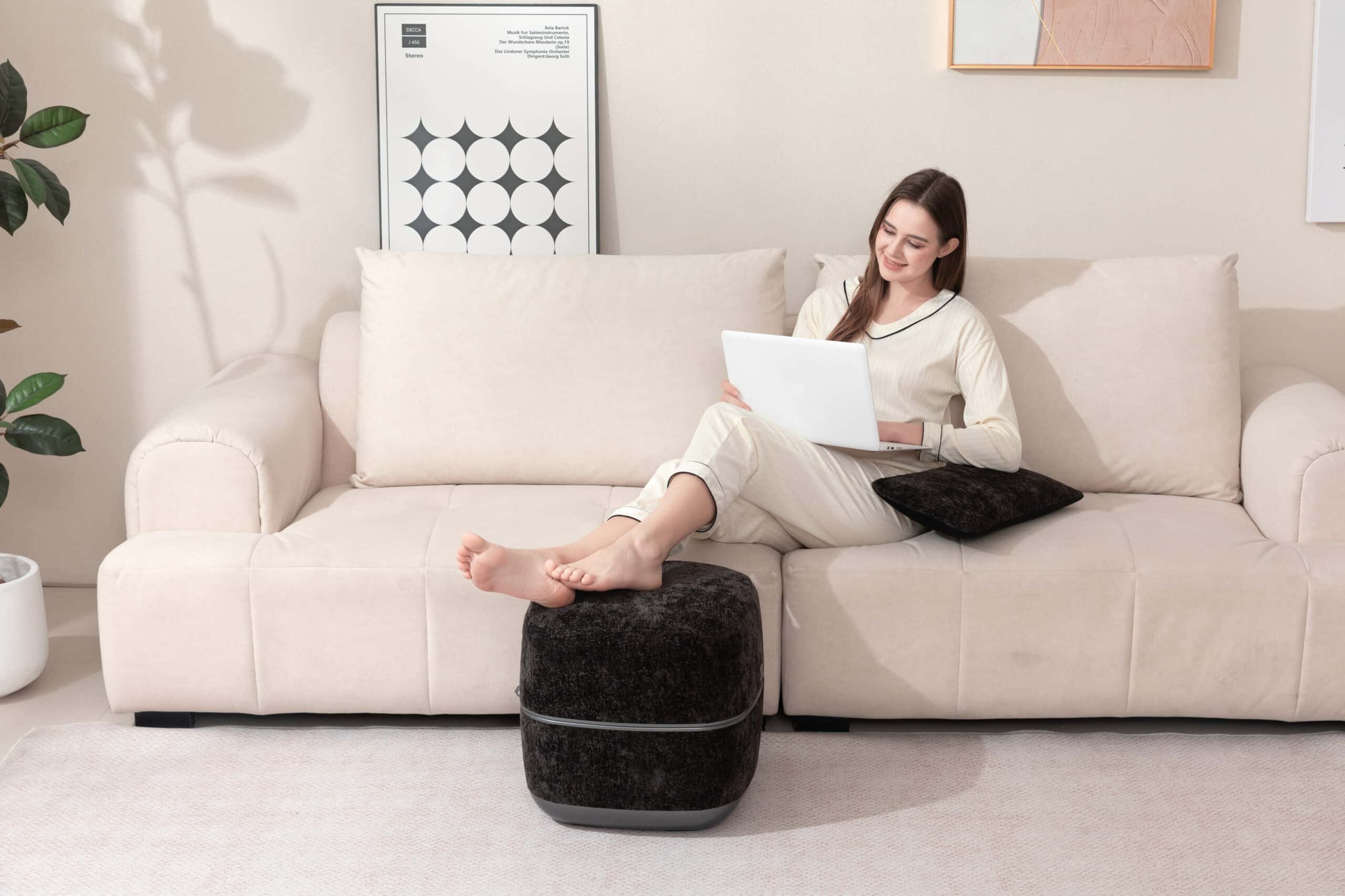 Kvinna i pyjamas använder en svart fotmassageapparat med öppet lock, sittandes i en ljus soffa och arbetar på datorn i ett harmoniskt hem.